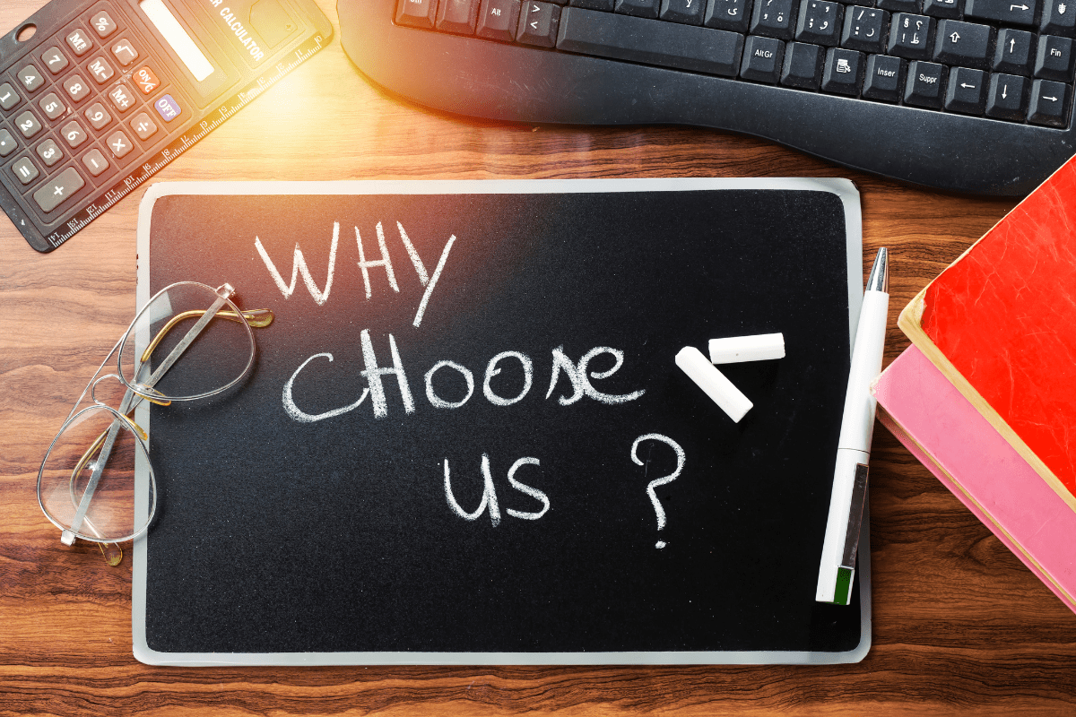 A small chalkboard on a wooden desk with the handwritten words “WHY CHOOSE US” surrounded by white chalk, a pen, eyeglasses, a calculator, a keyboard, and red and pink notebooks— A small chalkboard on a wooden desk with the handwritten words “WHY CHOOSE US” surrounded by white chalk, a pen, eyeglasses, a calculator, a keyboard, and red and pink notebooks—suggesting a business or academic setting.