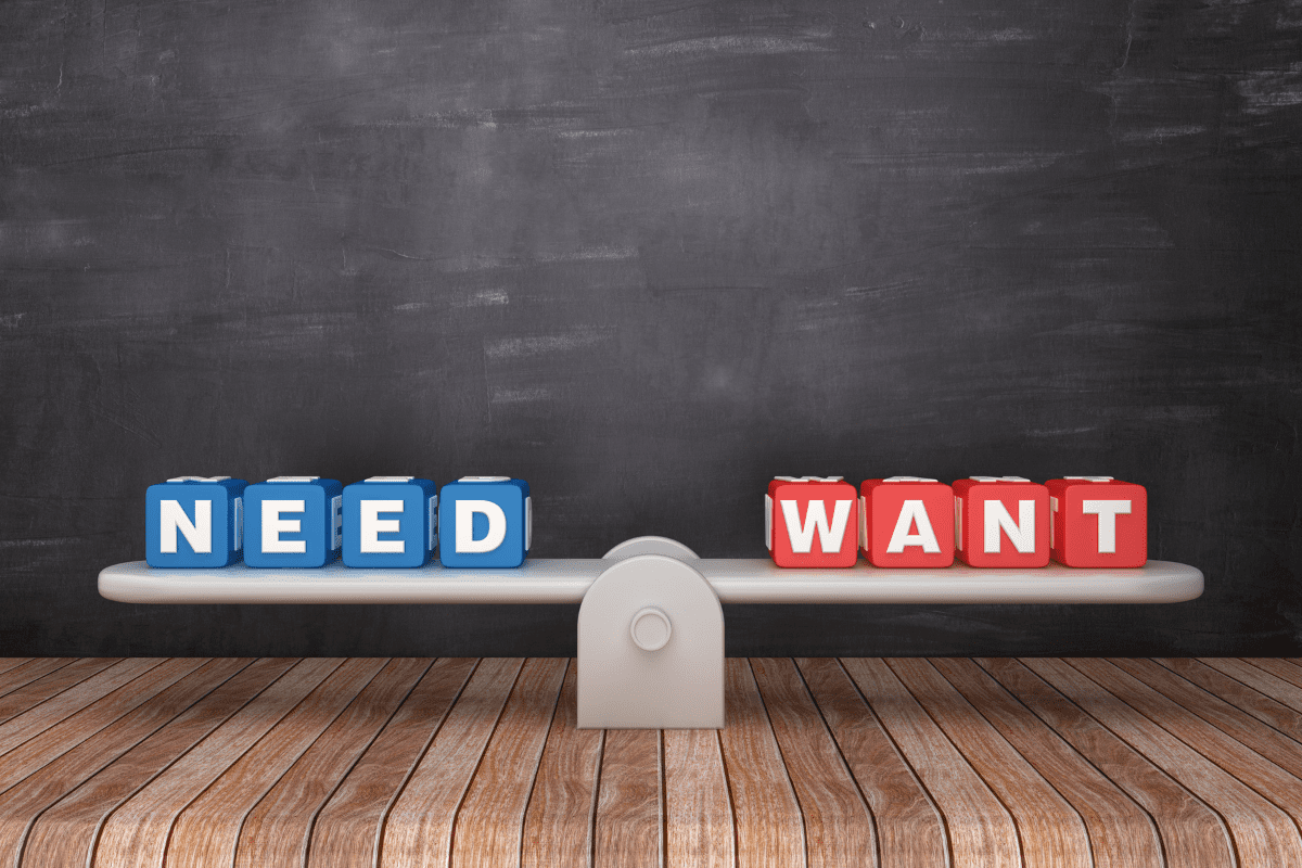 need want A balancing scale on a wooden surface with blue lettered cubes spelling “NEED” on the left side and red cubes spelling “WANT” on the right side, set against a chalkboard background—illustrating the contrast between needs and wants.